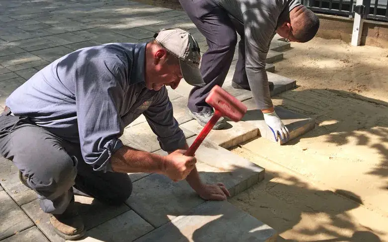 Two contractors repairing a cracked driveway by installing concrete pavers over a sand base.